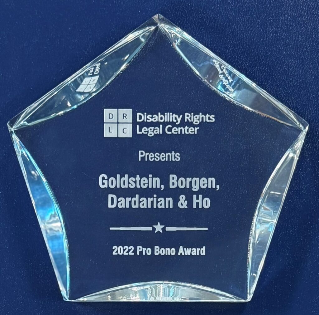 Disability Rights Legal Center, Pro Bono Award, 2022 Pentagon-shaped crystal award engraved with D R L C logo at top left with Disability Rights Legal Center written on the right side of the logo. Presents Goldstein, Borgen, Dardarian & Ho the 2022 Pro Bono Award. The award is clear, faceted edges reflect light against a dark blue background.