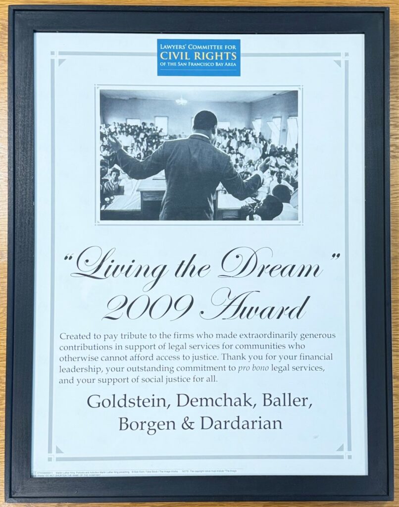 Lawyer’s Committee of San Francisco, Living the Dream Award, 2009 Framed certificate is displayed in a dark wood or black frame on a wooden surface. At the top is a blue Lawyers’ Committee for Civil Rights of the San Francisco Bay Area logo. Below it is a black-and-white photograph showing a man seen from behind speaking or addressing a large seated audience in a crowded room. The center of the certificate features the large script title, “Living the Dream,” followed by “2009 Award.” Beneath the title, the certificate states, "Created to pay tribute to the firms who made extraordinarily generous contributions in support of legal services for communities who otherwise cannot afford access to justice. Thank you for your financial leadership, your outstanding commitment to pro bono legal services, and your support of social justice for all." "Goldstein, Demchak, Baller, Borgen & Dardarian's name is written beneath that. The certificate has a grey decorative border with corner accents.
