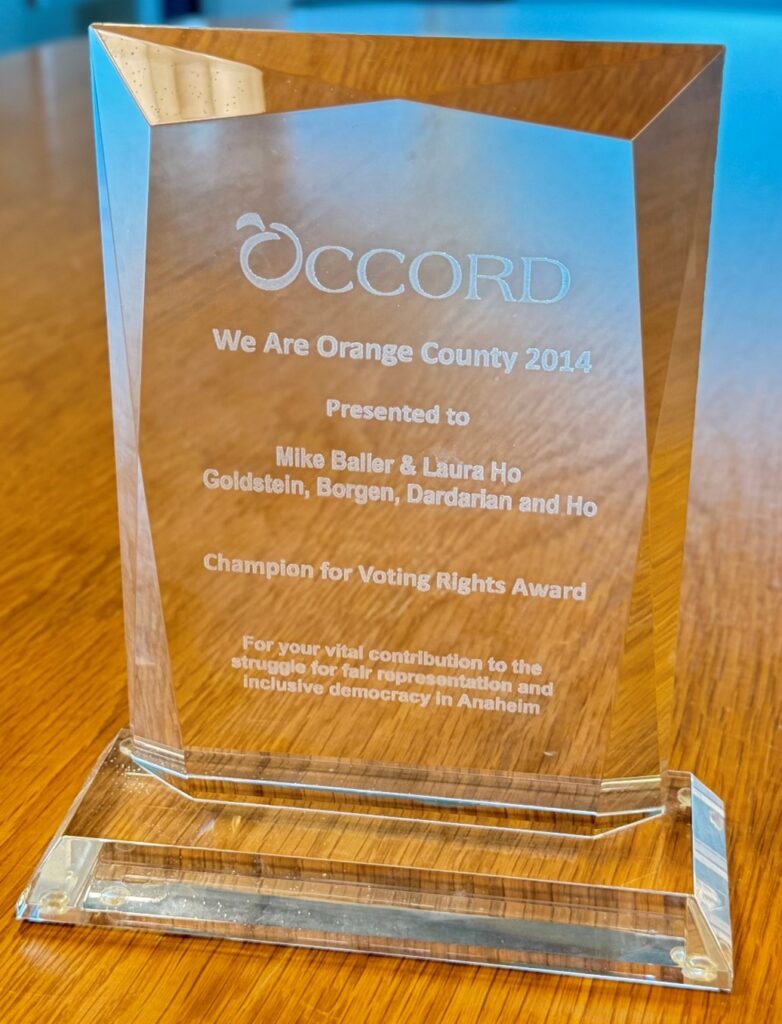 OCCORD, We are Orange County, 2014 Photograph of a clear glass award trophy with angled edges placed on a wooden table. Trophy is engraved with "OCCORD We Are Orange County 2014" presented to Mike Beller & Laura Ho from Goldstein, Borgen, Dardarian and Ho Champion for Voting Rights Award for your vital contribution to the struggle for fair representation and inclusive democracy in Anaheim.